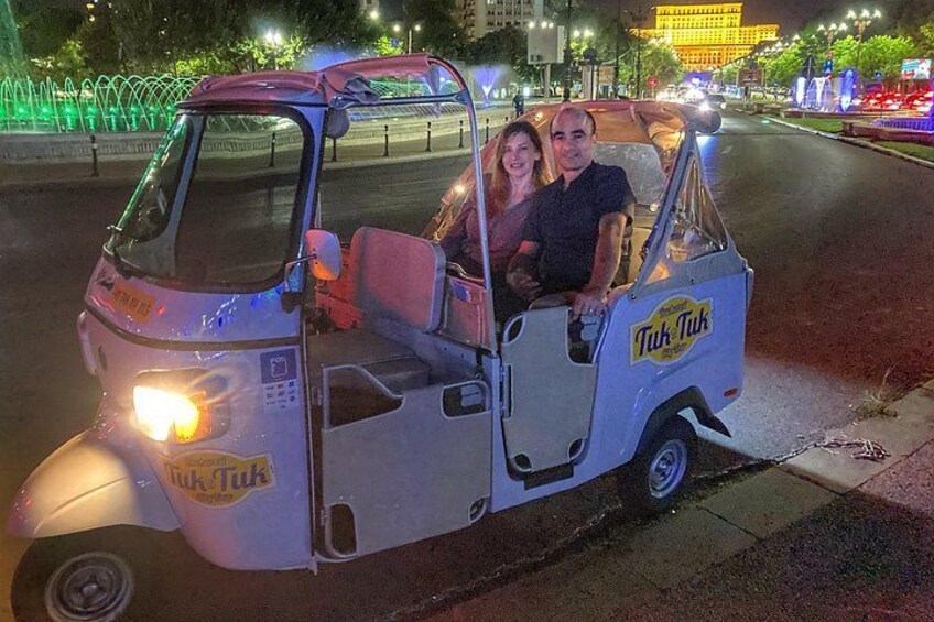 Een tuk-tuk met twee passagiers, een man en een vrouw, op een stadstraat 's nachts. 2