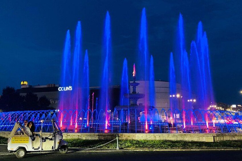 Een nachtscène met een fonteinshow met blauwe en roze verlichting, een McDonald's bord en een voertuig op de voorgrond. 4