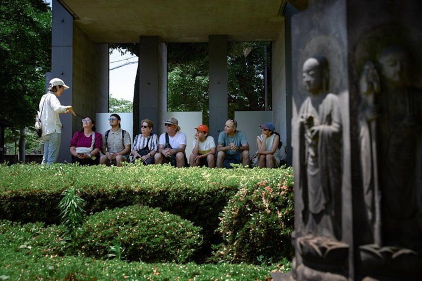 Eine Gruppe von Menschen sitzt auf einer Bank unter einem Schutzdach, im Vordergrund steht eine Statue. 5
