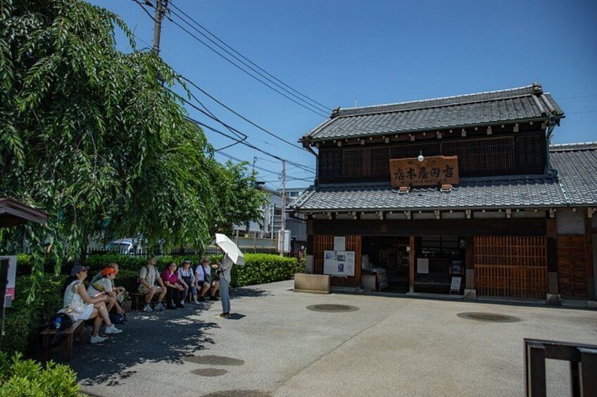 Ein traditionelles japanisches Gebäude mit einem Schild, Personen, die auf Bänken sitzen, und einem Baum im Vordergrund. 2