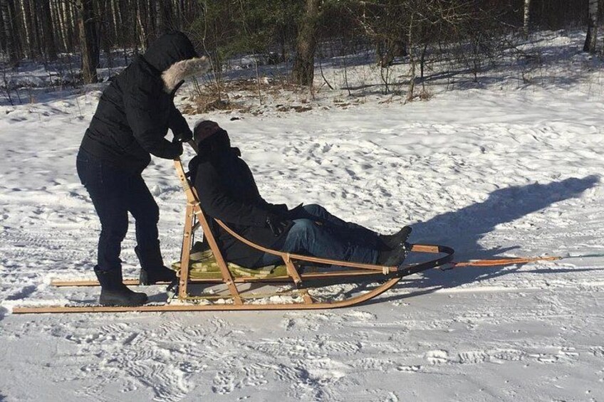 A person in a black jacket and hat pushing a sled with a person seated in it on a snowy ground. 1