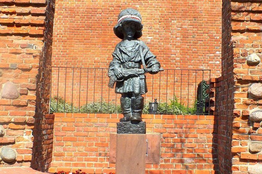 A statue of a child in a hat and coat, standing on a pedestal, with a brick wall and metal fence in the background. 4