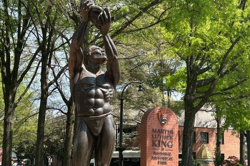 A bronze statue of a muscular man holding a basketball, with a red brick structure labeled 'Martin Luther King Jr. National Historical Park' in the background. 5