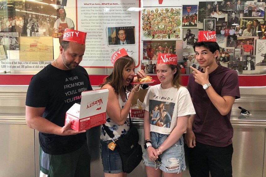 People wearing hats, holding food, and standing in a restaurant with a menu board in the background. 1