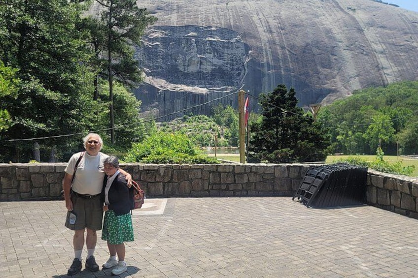 Two people standing in front of a large rock formation with a paved walkway and greenery. 2