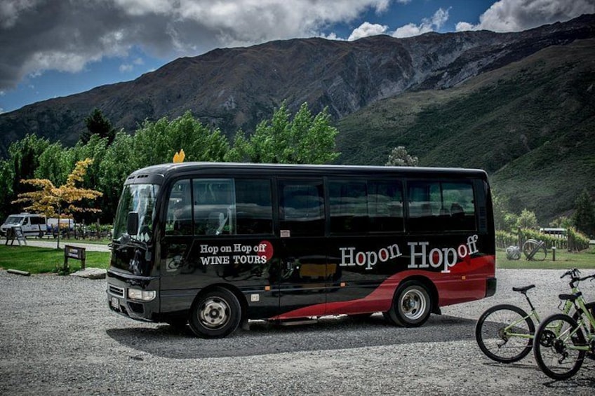 Un autobus noir et rouge, sur lequel on peut lire « Visites guidées des vignobles, montées et descentes à volonté », est garé sur un sol recouvert de gravier. On aperçoit une montagne et des arbres en arrière-plan. 1