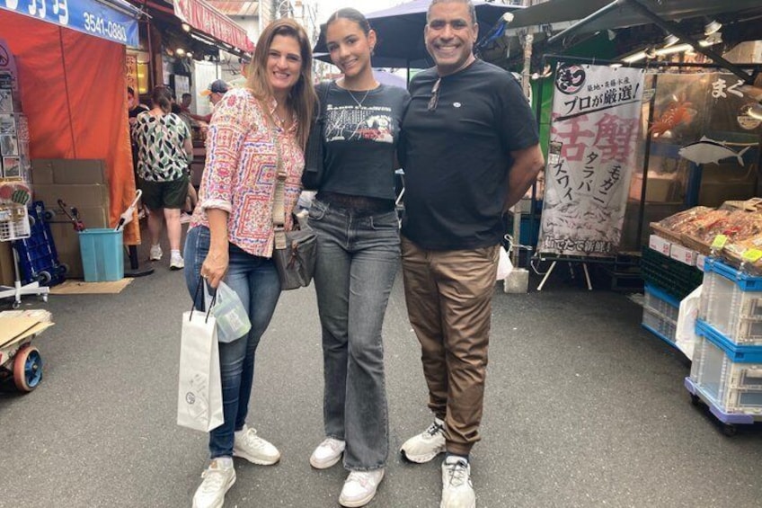 Tres personas están de pie en un mercado al aire libre, con varios puestos y mercancías al fondo. 4