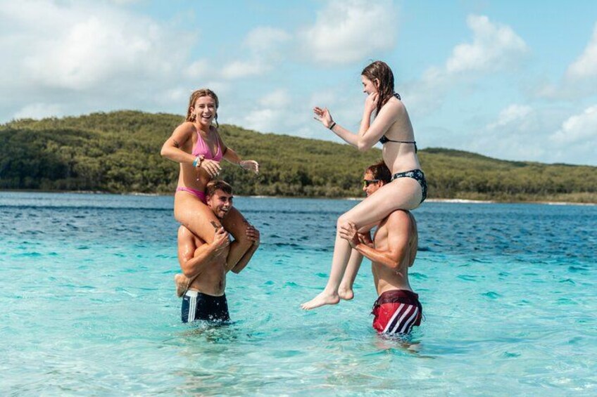 Trois personnes dans une étendue d’eau, dont une porte une autre sur ses épaules, et une colline boisée en arrière-plan. 2
