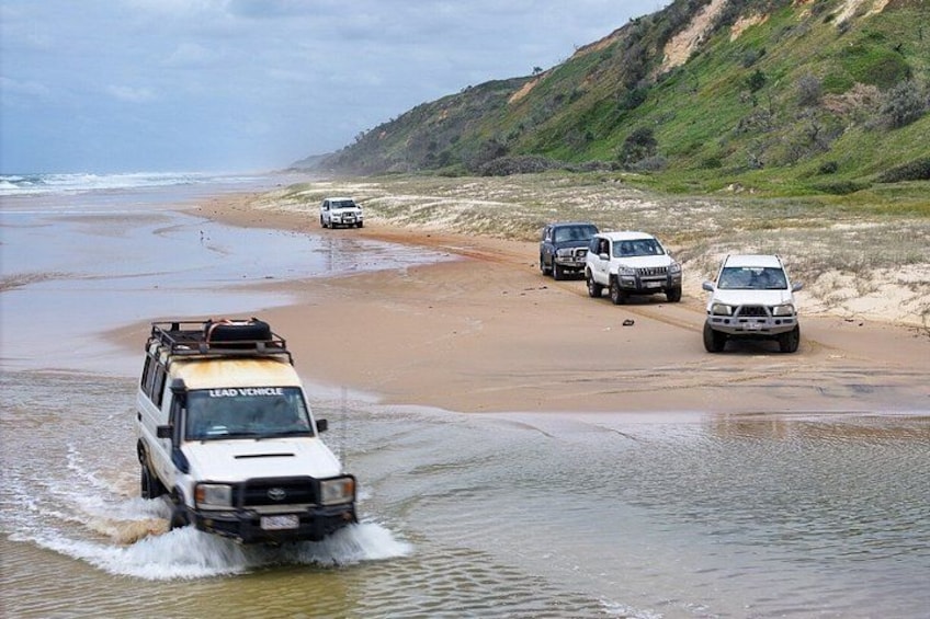 A white SUV driving through shallow water on a beach with other vehicles parked nearby. 2