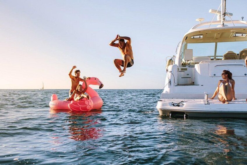 En person hoppar från en rosa uppblåsbar flamingofigur ner i vattnet. I bakgrunden syns en lyxyacht och en segelbåt. 2