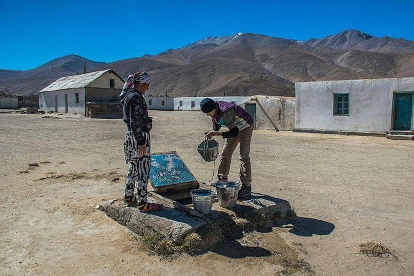 Two people in a desert setting with mountains in the background, one standing and one bending over a container. 1