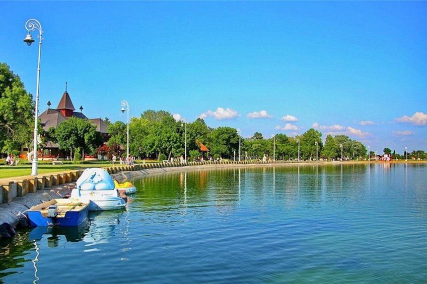 A calm lake with boats docked, a park with trees and a building with a tower, and a clear blue sky. 2
