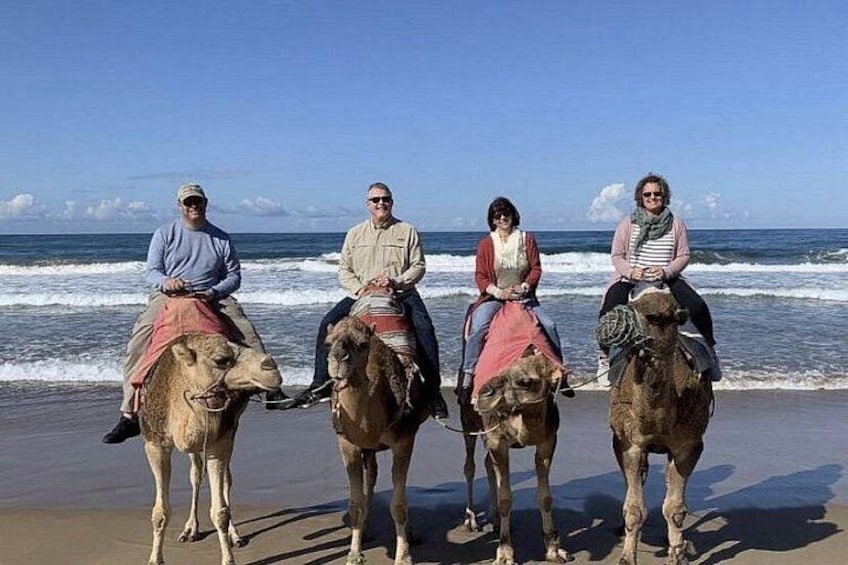 Four people riding camels on a beach with the ocean in the background. 3