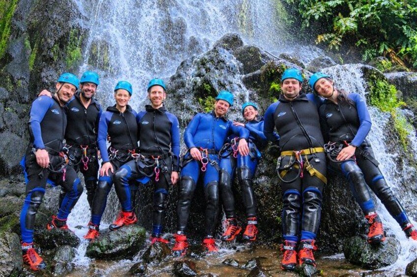 Een groep mensen in blauwe neopreenpakken en helmen poseren voor een waterval. 2