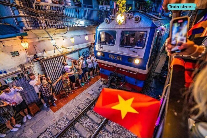 A train with the number 019F-928 on the front, passing by a crowd of people on a platform, with a flag of Vietnam in the foreground. 4