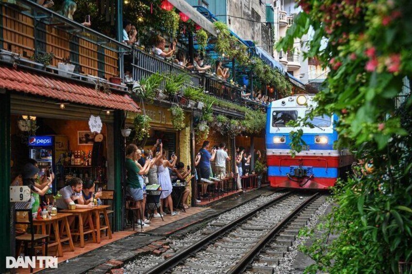 A train passing through a bustling urban area with people gathered on the sidewalk, a restaurant with outdoor seating, and greenery on the buildings. 3