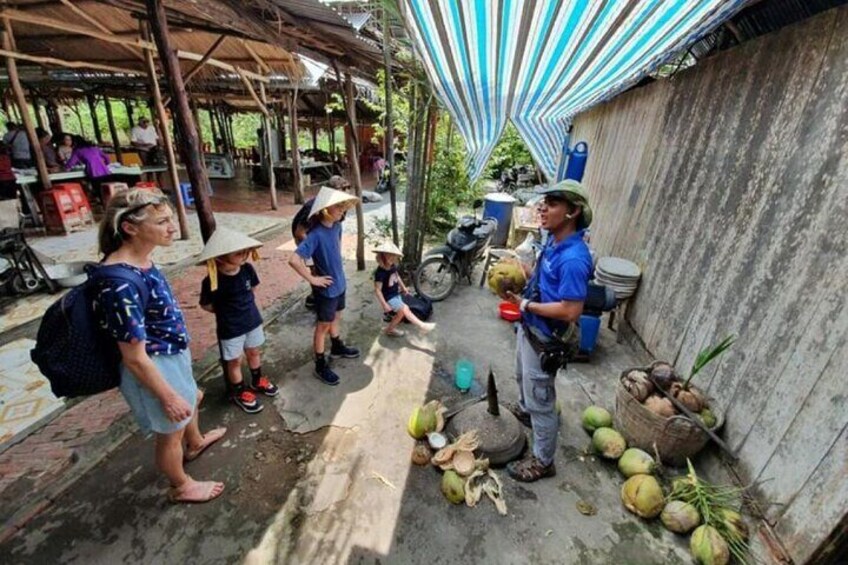 A group of people outdoors, some wearing hats, standing near a wall with coconuts and a motorcycle. 3