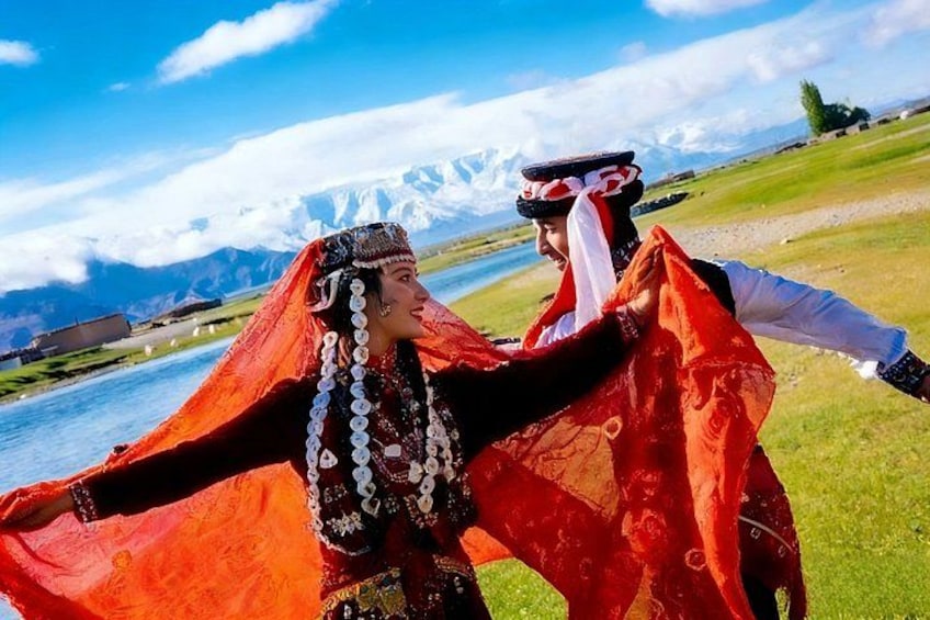 Two people in traditional attire with a mountainous landscape and a body of water in the background. 1