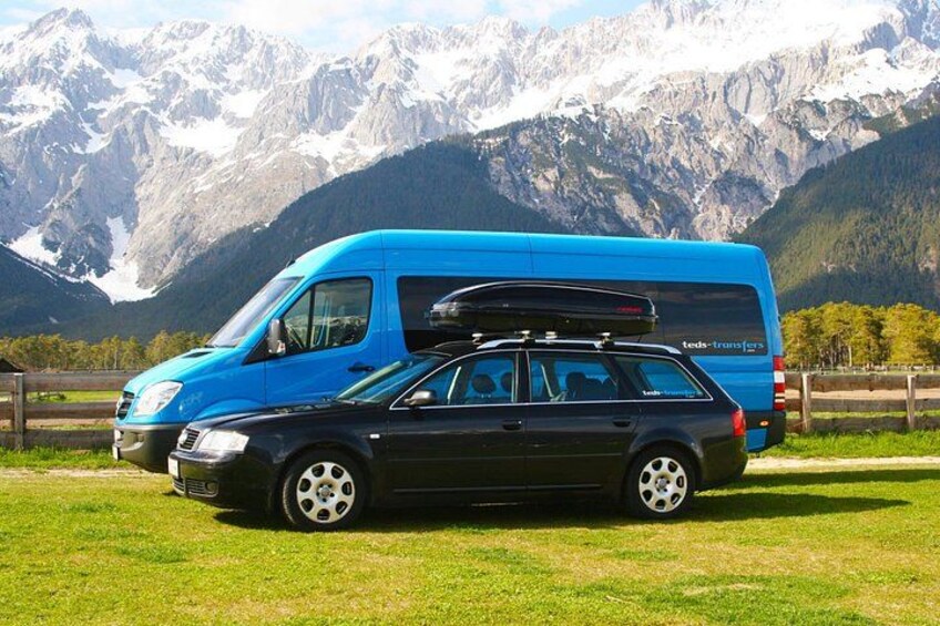 Un fourgon bleu avec un porte-bagages sur le toit et une voiture noire sont stationnés dans un champ herbeux, avec des montagnes en arrière-plan. 1