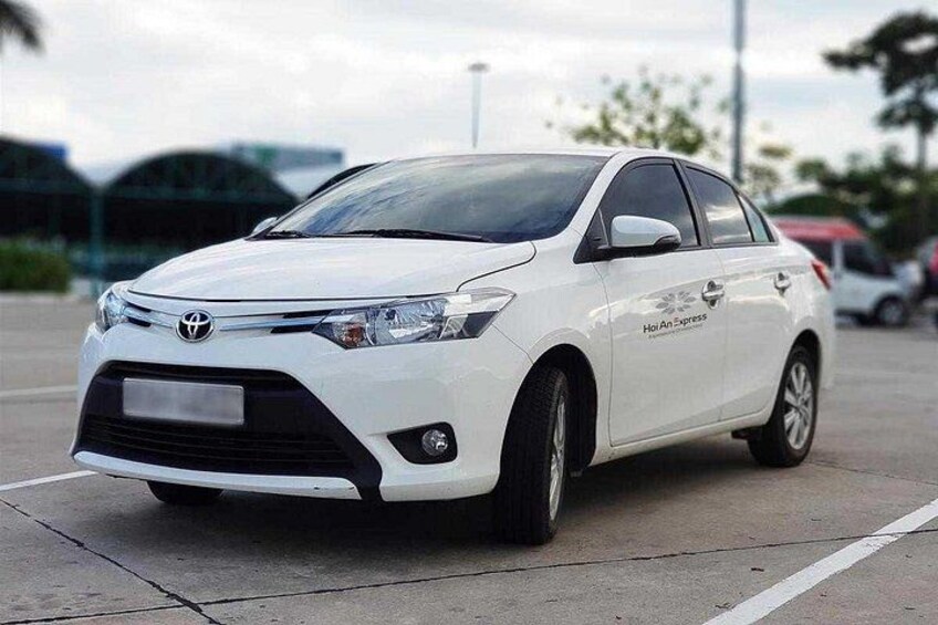 Une berline Toyota blanche est garée sur une surface en béton, avec un arrière-plan flou composé d’arbres et de bâtiments. 4