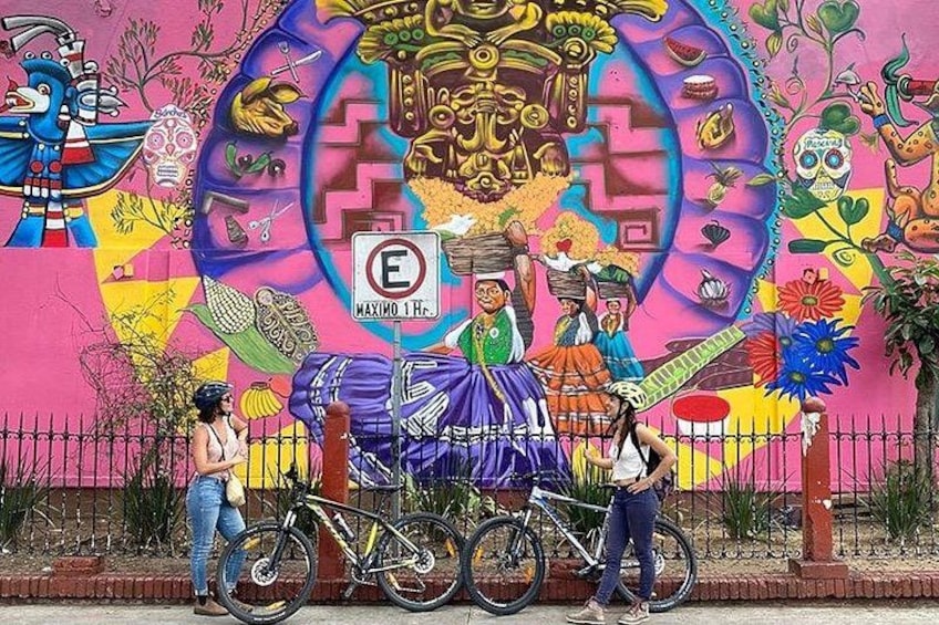 Et farverigt mural med kulturelle motiver, en cykel og en person ved hegnet. 1