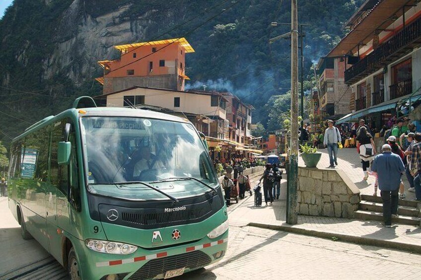 A green bus on a street with people walking and buildings in the background. 1