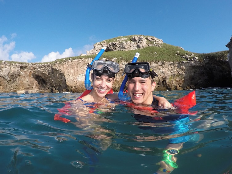 Dos personas haciendo snorkel en aguas azules y cristalinas, con acantilados rocosos y vegetación al fondo. 1