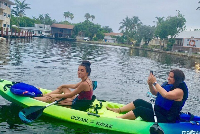 Dos personas en kayak en un río tranquilo, con edificios y palmeras al fondo. 1