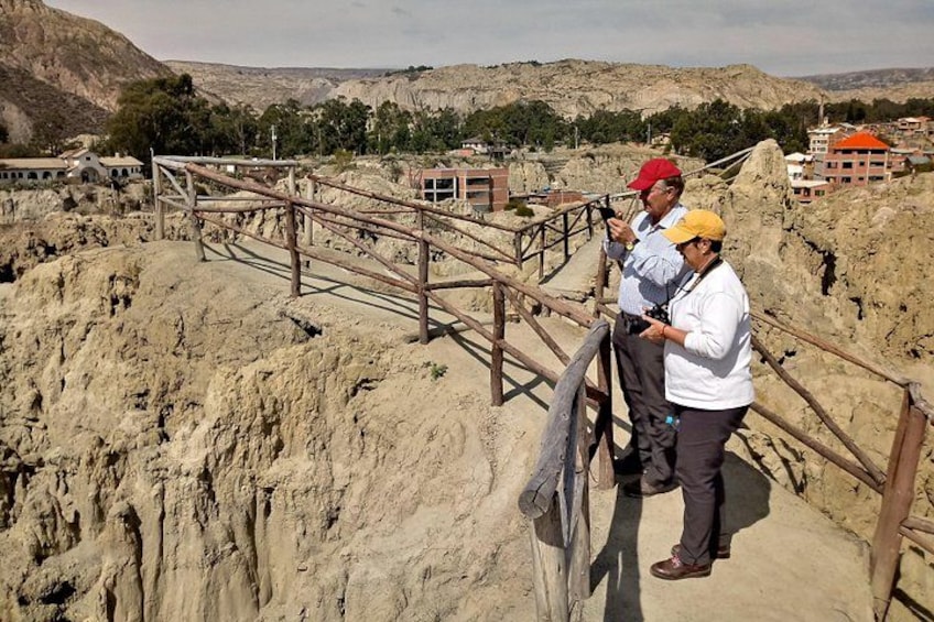 Two people standing on a wooden staircase in a rocky area with a cityscape in the background. 5