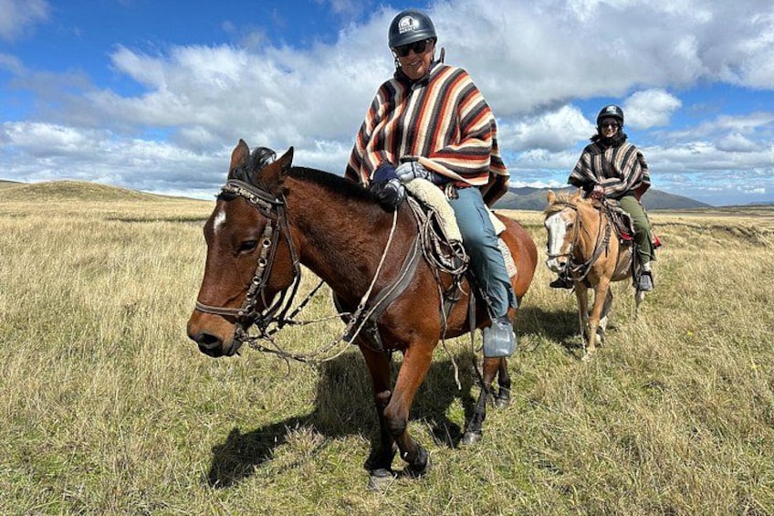 Two people riding horses in a grassy field with a mountainous background. 1