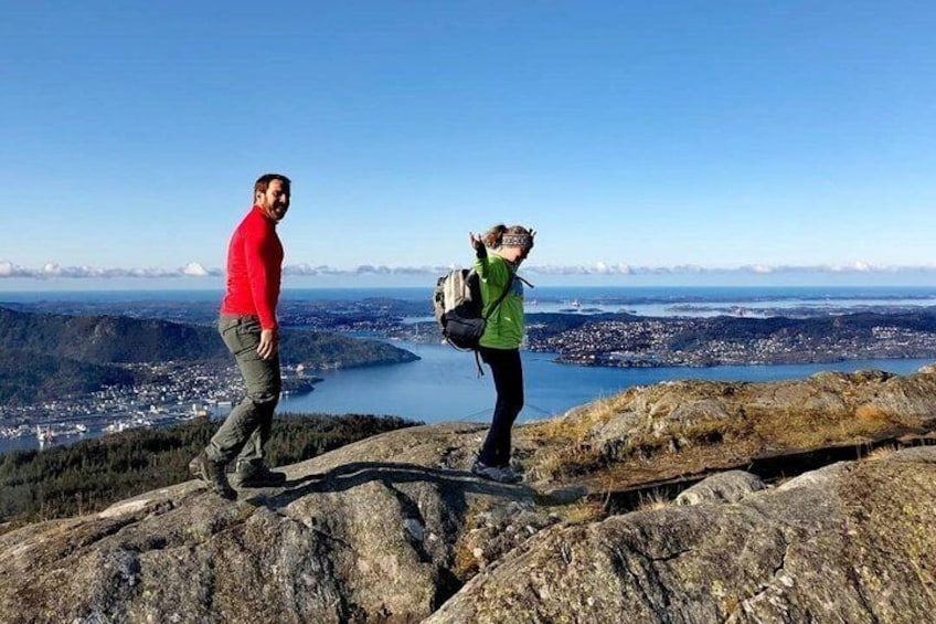 Two people on a rocky outcrop overlooking a valley with a lake and distant cityscape. 2