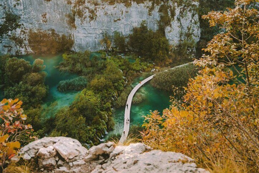 A narrow bridge over a clear green river, surrounded by dense foliage and a rocky cliff. 2