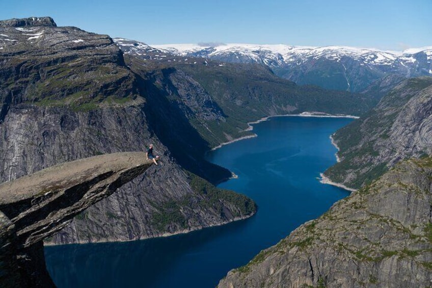 Une personne se tient au bord d’une falaise, surplombant un fjord d’un bleu profond, entouré de montagnes escarpées et de sommets enneigés. 2