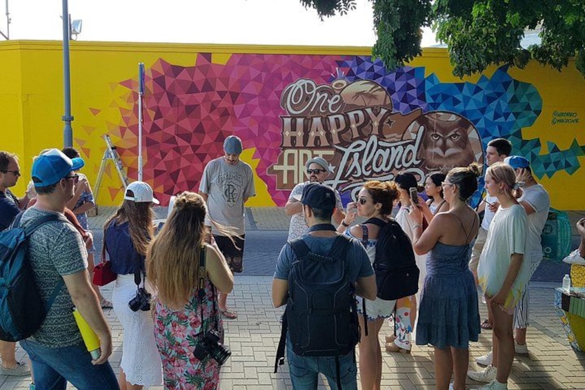 A group of people gathered in front of a colorful mural with the text 'One Happy Island.' 5