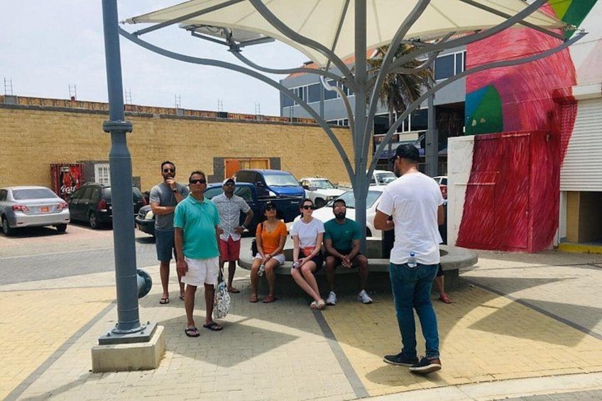A group of people sitting on a bench under a shaded area, with a building and parked cars in the background. 3