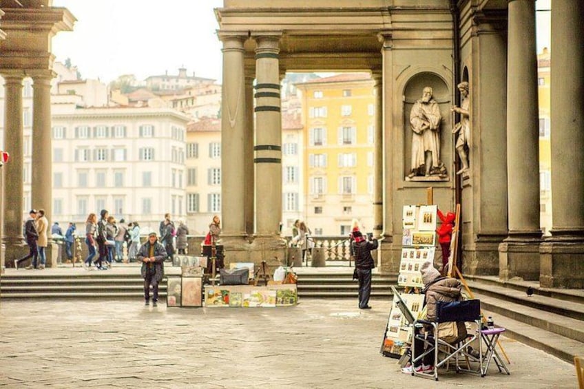 A public square with classical architecture, people walking, and a street vendor with art supplies. 2