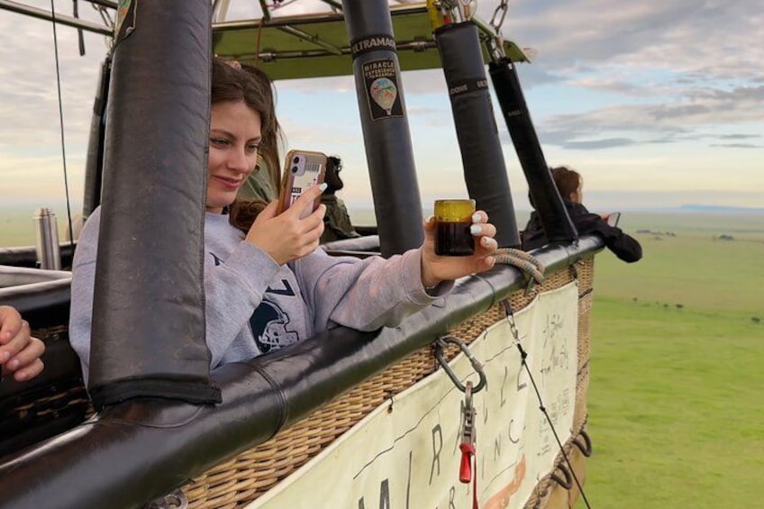 Een persoon in de gondel van een luchtballon, vasthoudend een smartphone en een drankje. 5