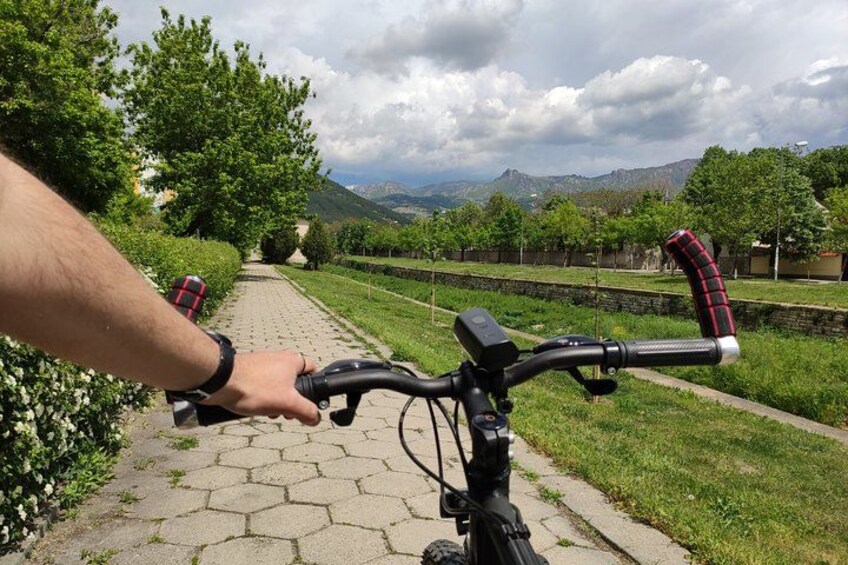 A paved path with a hand on a bicycle handlebar, surrounded by greenery and distant mountains. 3