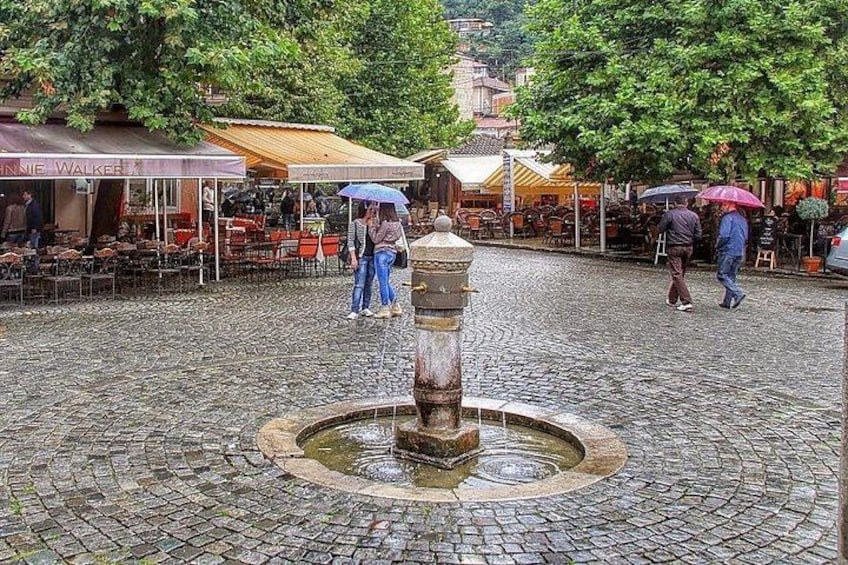 A cobblestone square with a central fountain, outdoor seating at cafes, and people walking with umbrellas. 1