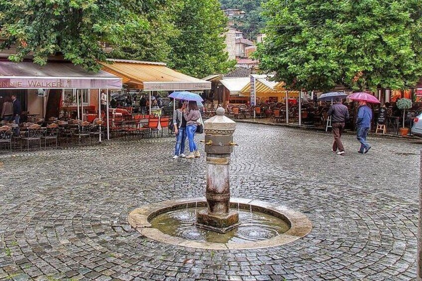 A cobblestone square with a central fountain, outdoor seating areas, and people walking with umbrellas. 4
