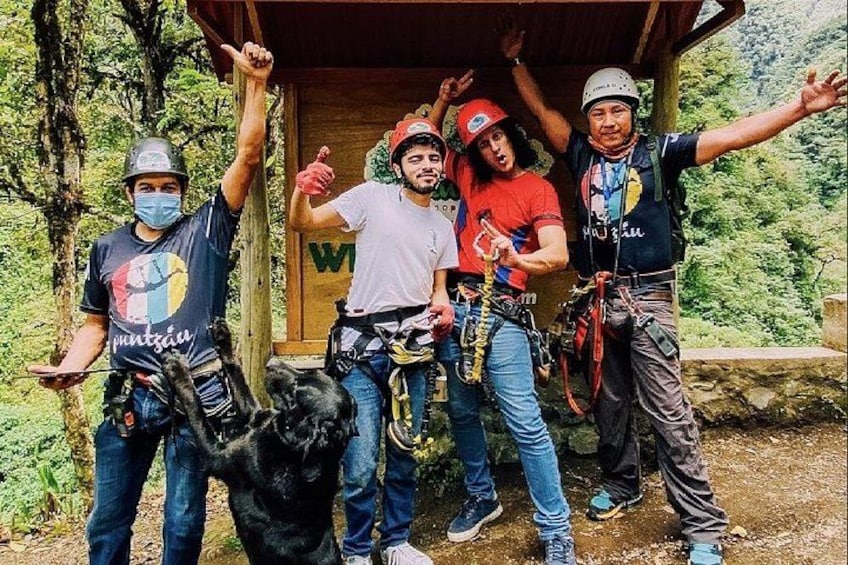 Un grupo de personas con cascos y arneses, de pie en un área boscosa con una estructura de madera al fondo. 4