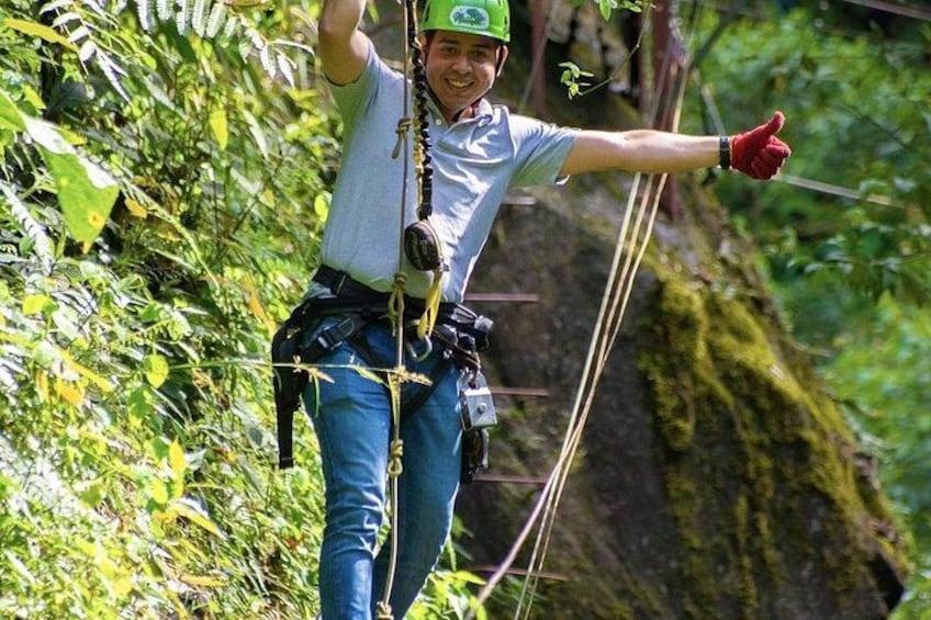 Una persona deslizándose en un tirolina, usando casco y guantes, con follaje y un risco rocoso al fondo. 5