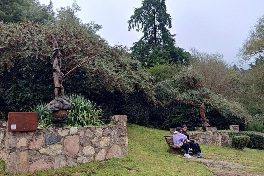 Una estatua sobre un pedestal de piedra, un área con césped y una persona sentada en un banco. 4