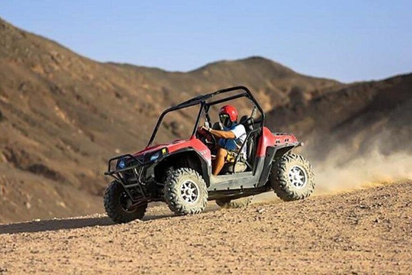 A red ATV with two people, one wearing a helmet, driving on a dirt road with a mountainous background. 2
