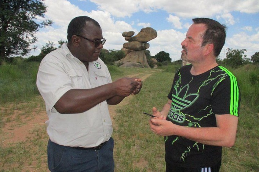 Two men outdoors, one wearing a white shirt and the other in a black Adidas t-shirt, standing on a grassy area with a large rock formation in the background. 2