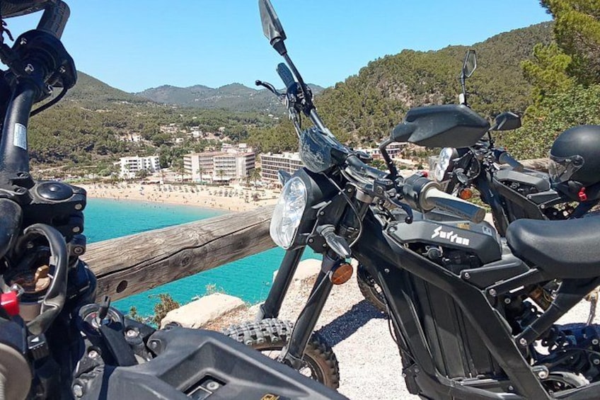 Una motocicleta estacionada junto a una baranda de madera con vista a una playa y edificios al fondo. 4