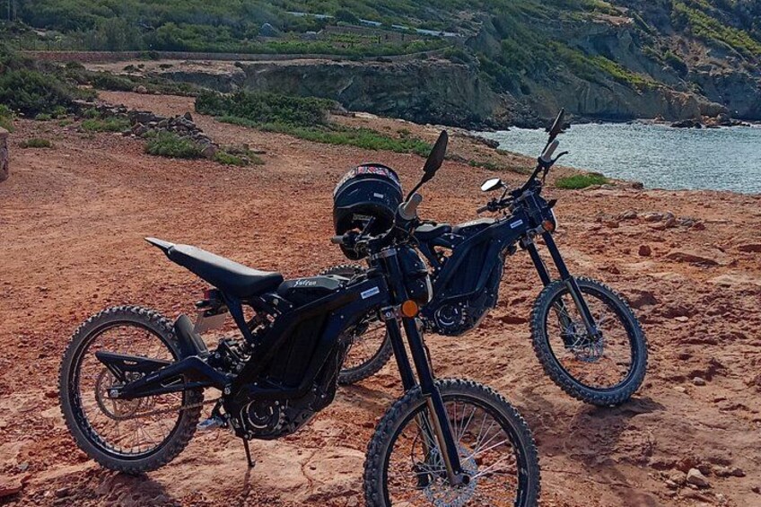 Una moto de cross estacionada en una playa rocosa, con una masa de agua y vegetación al fondo. 2
