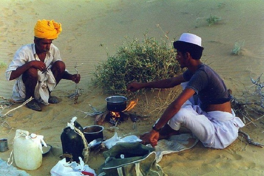 Two people cooking over an open fire in a desert setting with cooking utensils and a pot on the ground. 2