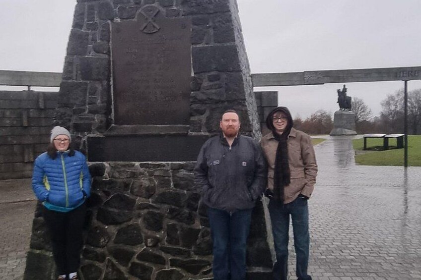 Trois personnes se tiennent devant un monument de pierre orné d’inscriptions, sous un pont. Une statue est visible en arrière-plan. 4