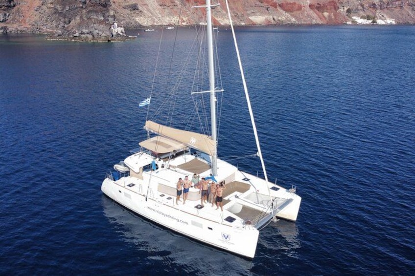 A white sailboat with a covered deck and passengers, anchored in calm blue waters with rocky cliffs in the background. 3